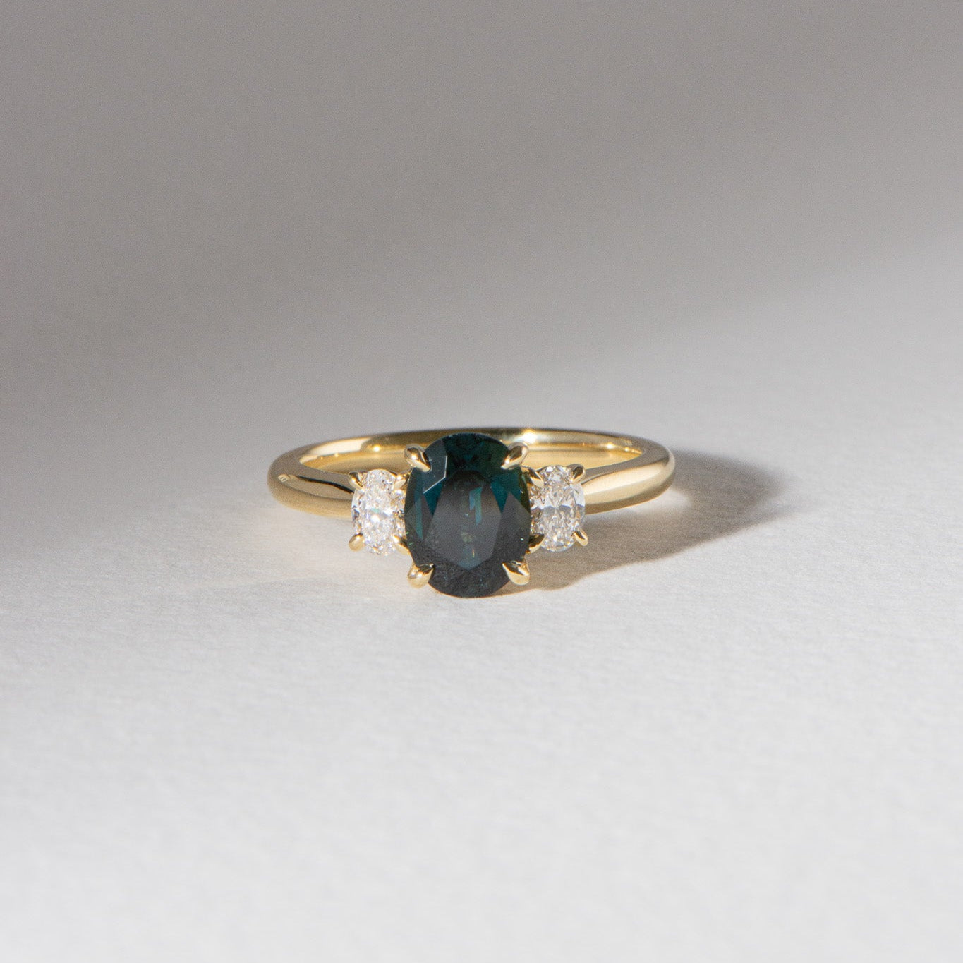 Australian blueish teal sapphire engagement ring with 1.78ct centre sapphire, 0.29ct oval lab diamonds, set in 18k yellow gold cathedral three-stone setting.