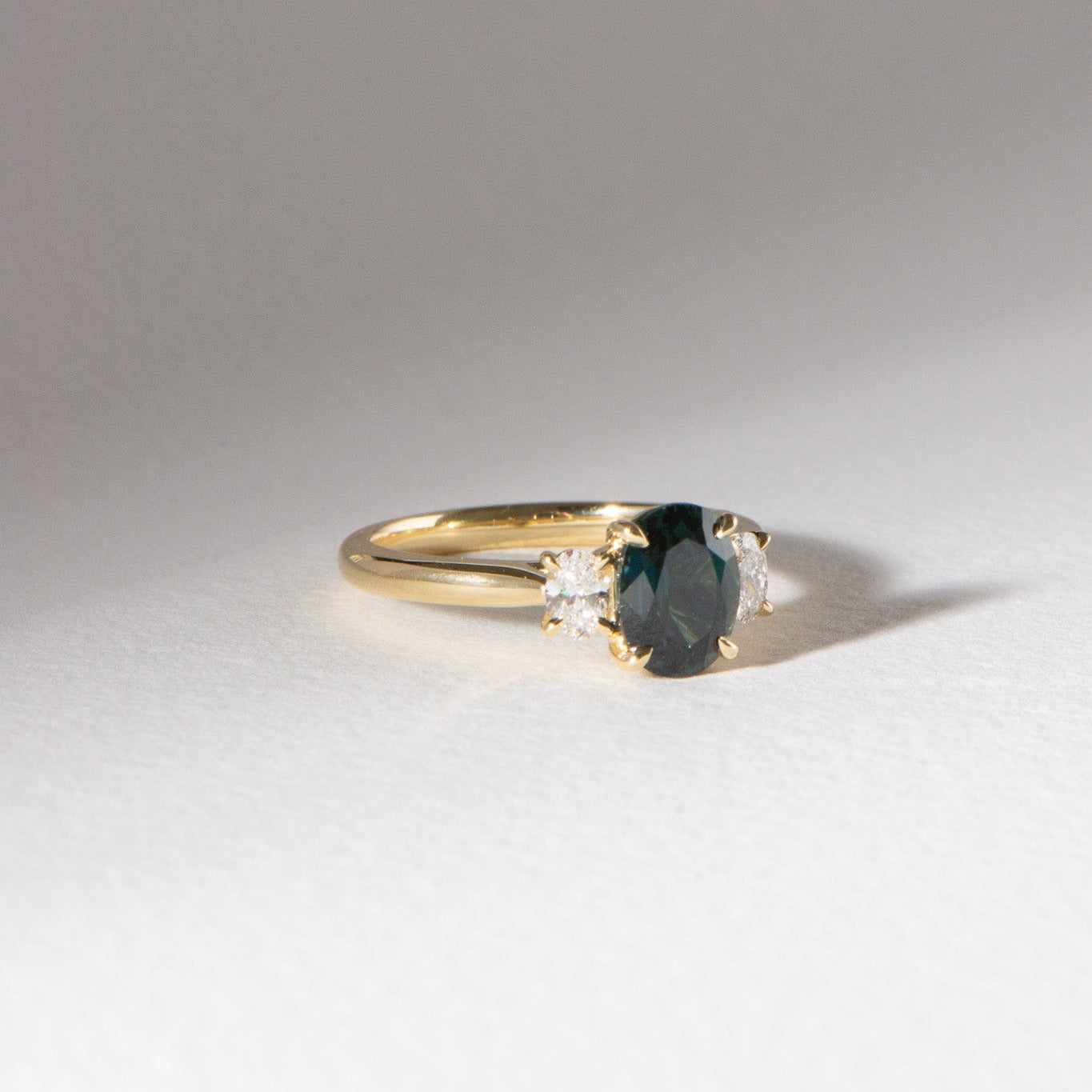 Australian blueish teal sapphire engagement ring with 1.78ct centre sapphire, 0.29ct oval lab diamonds, set in 18k yellow gold cathedral three-stone setting.
